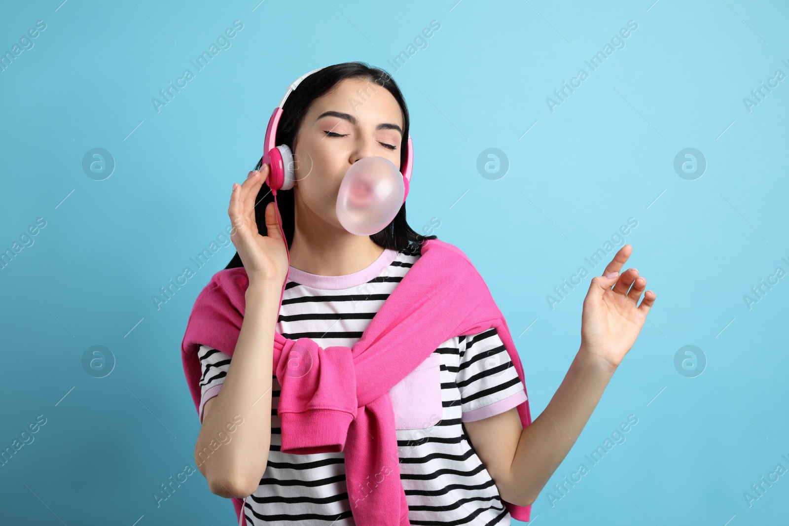 Photo of Fashionable young woman with headphones blowing bubblegum on light blue background