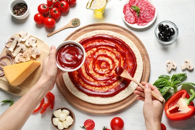 Woman spreading tomato sauce onto pizza crust and ingredients on white wooden table, top view