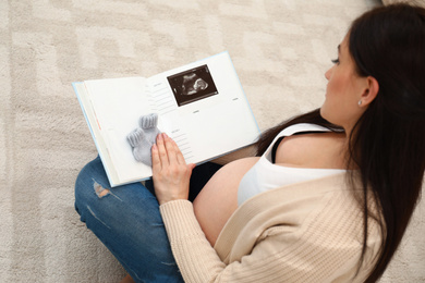 Photo of Beautiful pregnant woman holding memory book with socks and ultrasound photo of her baby at home, above view
