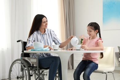 Mother in wheelchair and her daughter drinking tea at table indoors