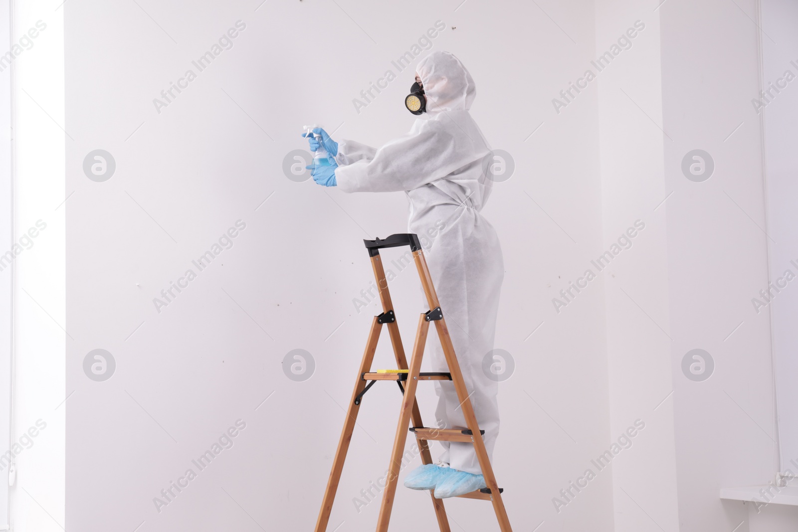 Photo of Woman in protective suit cleaning mold with sprayer on wall indoors