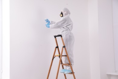 Woman in protective suit cleaning mold with sprayer on wall indoors