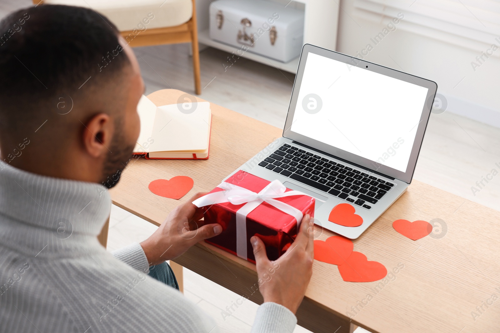 Photo of Valentine's day celebration in long distance relationship. Man holding gift box while having video chat with his girlfriend via laptop indoors