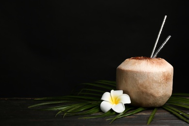 Fresh coconut with drinking straws and flower on wooden table against black background. Space for text