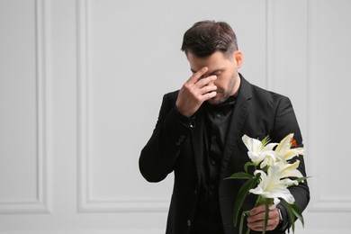 Photo of Sad man with lily flowers mourning near white wall, space for text. Funeral ceremony