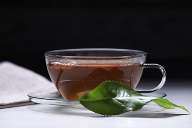 Photo of Aromatic hot tea in glass cup and leaf on white table against black background, closeup