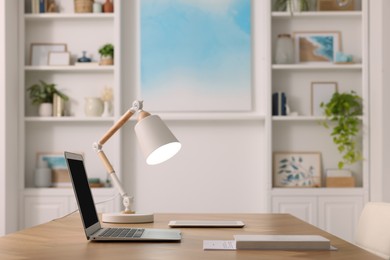 Photo of Interior design. Cosy workplace with laptop and lamp on wooden table near abstract picture between shelves