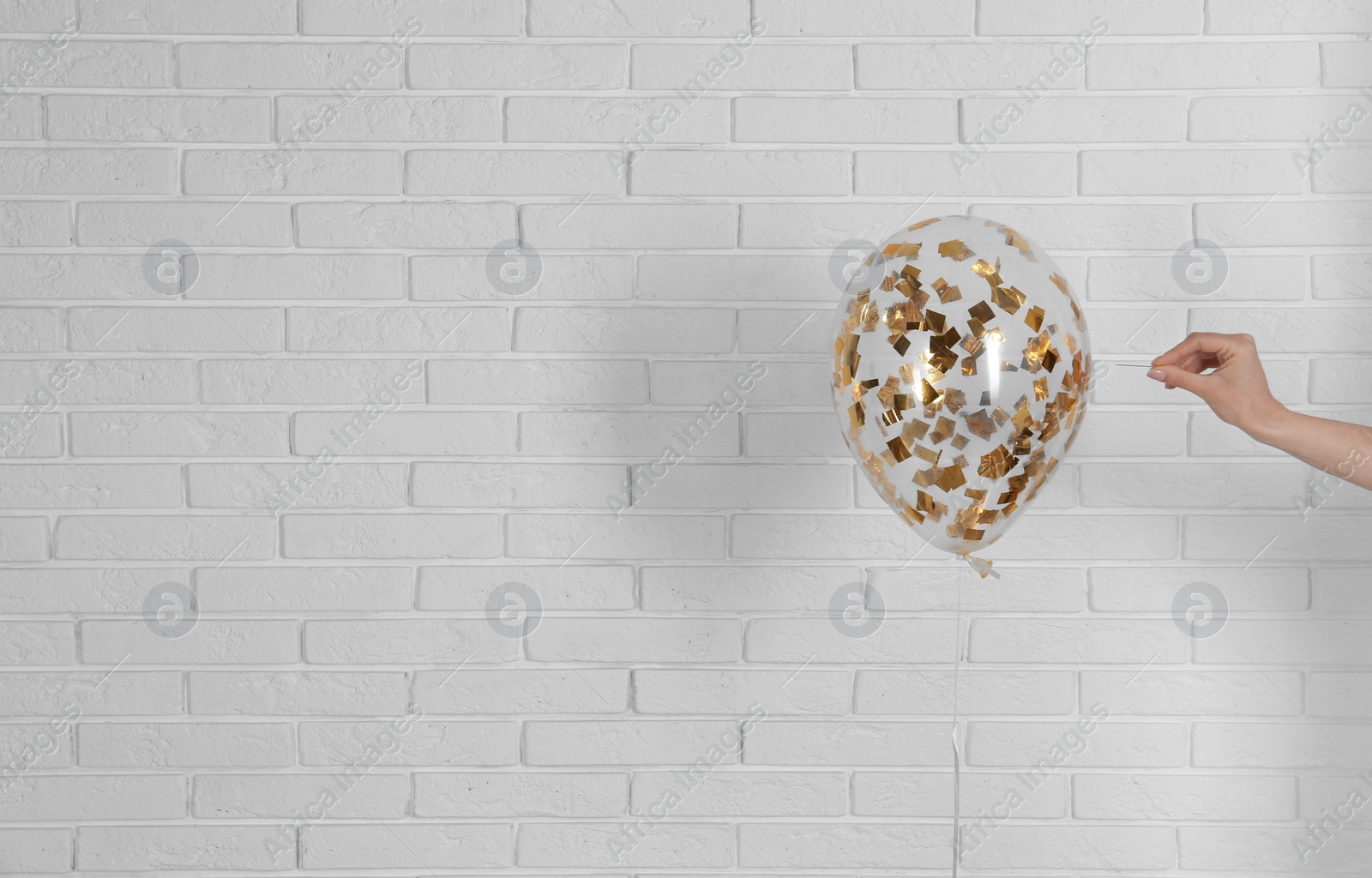 Photo of Young woman piercing bright balloon near brick wall, space for text