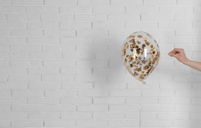 Young woman piercing bright balloon near brick wall, space for text