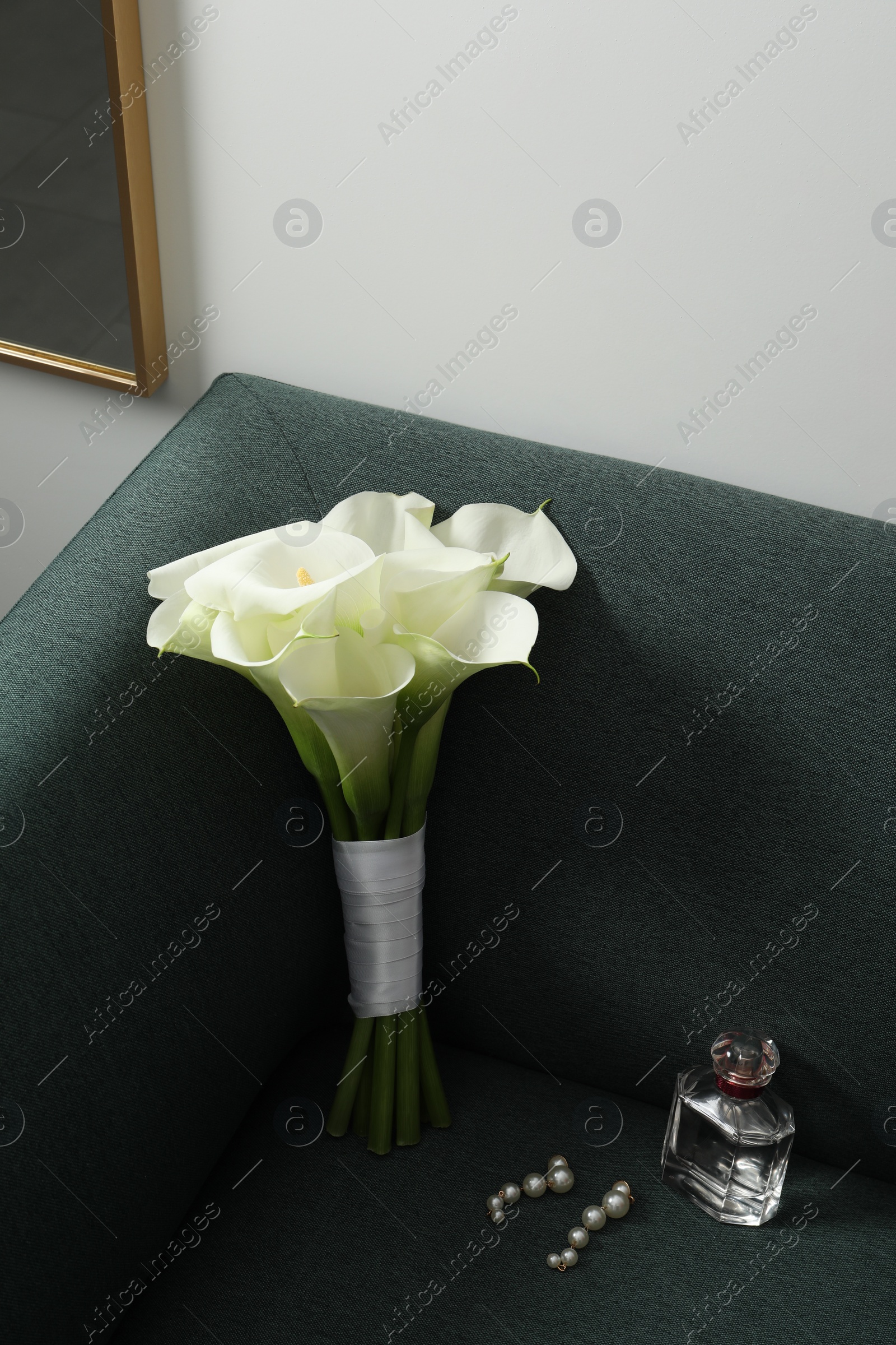 Photo of Beautiful calla lily flowers tied with ribbon, bottle of perfume and earrings on sofa indoors