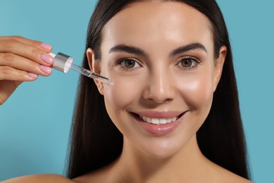 Photo of Beautiful young woman applying serum onto her face on light blue background, closeup