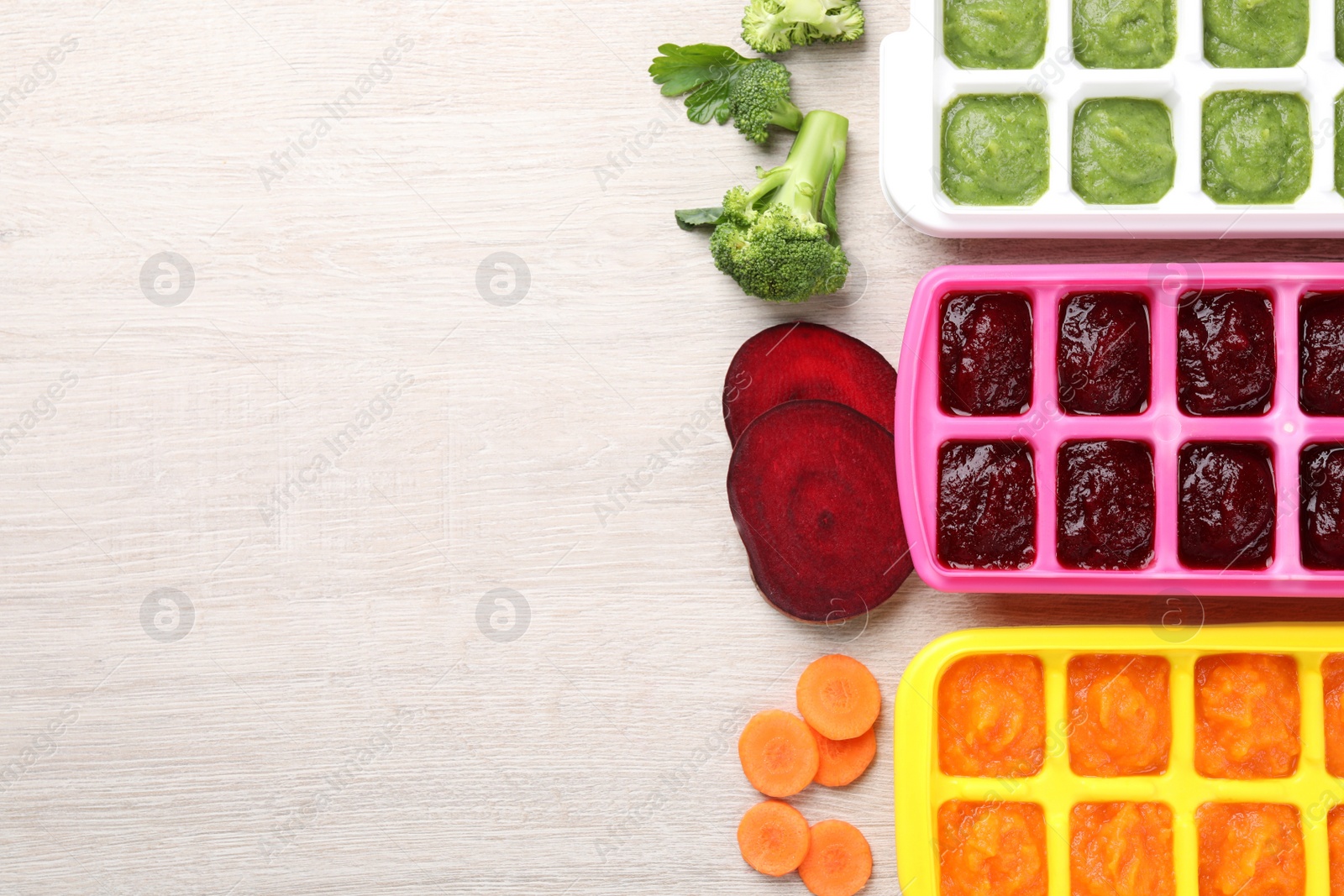 Photo of Different purees in ice cube trays ready for freezing and ingredients on white wooden table, flat lay. Space for text