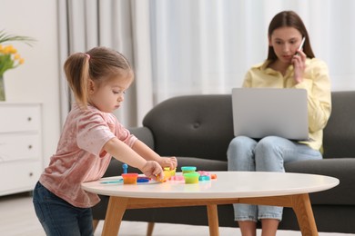 Photo of Woman working remotely at home. Mother talking on smartphone and using laptop while her daughter playing at desk, selective focus