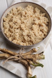 Photo of Bowl of tasty prepared horseradish and roots on white wooden table, above view