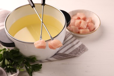 Photo of Fondue pot with oil, forks, raw meat pieces and parsley on white wooden table. Space for text