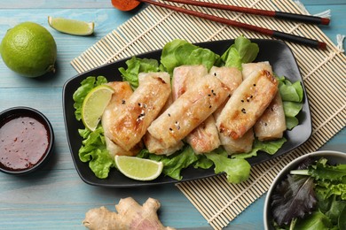 Photo of Tasty fried spring rolls, lettuce, lime and sauce on light blue wooden table, flat lay