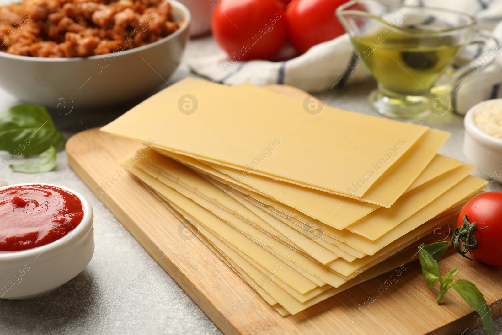 Photo of Cooking lasagna. Wooden board with pasta sheets and other products on grey textured table, closeup