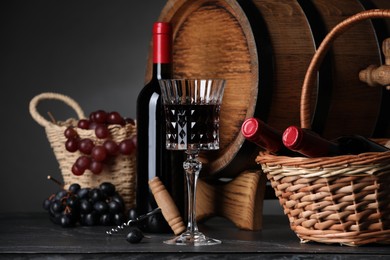 Glass of red wine, bottles, wicker basket, grapes and wooden barrel on black table