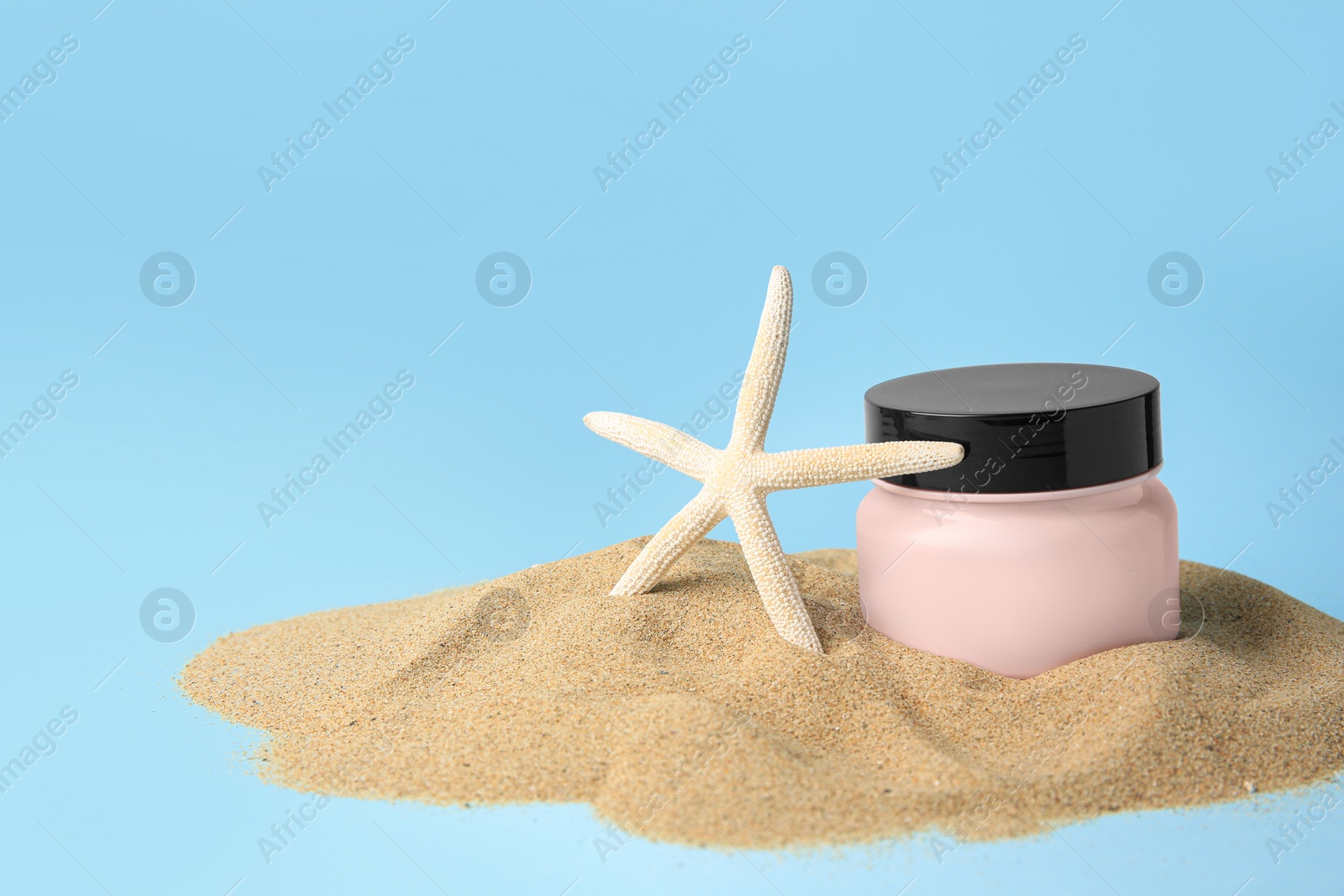 Photo of Jar with cream and starfish on sand against light blue background, space for text. Cosmetic product