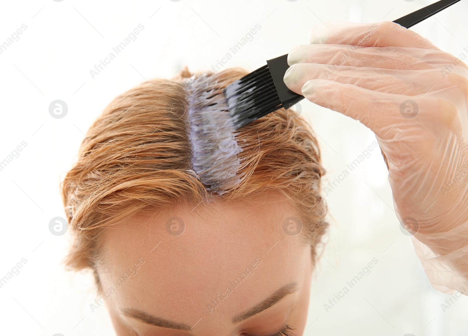 Photo of Young woman applying hair dye on roots against light background, closeup