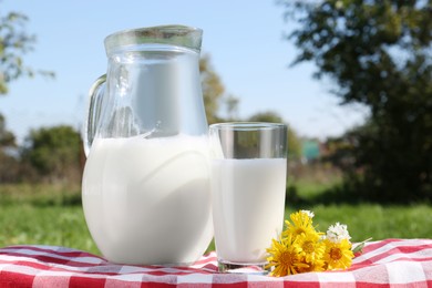 Photo of Jug and glass of fresh milk and flowers on checkered blanket outdoors