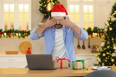 Celebrating Christmas online with exchanged by mail presents. Man in Santa hat covering eyes before opening gift box during video call on laptop at home