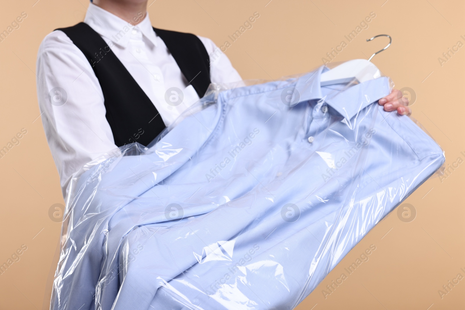 Photo of Dry-cleaning service. Woman holding shirt in plastic bag on beige background, closeup