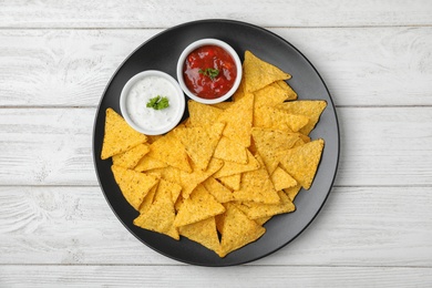 Photo of Black plate with tasty Mexican nachos chips and sauces on white wooden table, top view