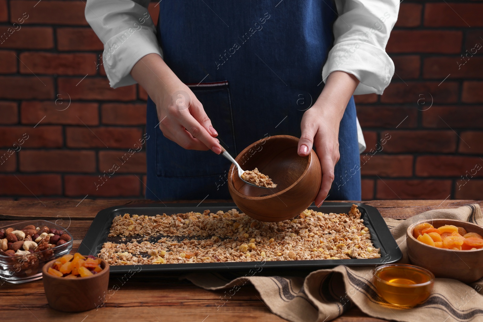 Photo of Woman putting granola from baking tray into bowl at wooden table, closeup