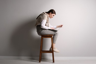 Photo of Young woman with bad posture using laptop while sitting on stool near light grey wall indoors
