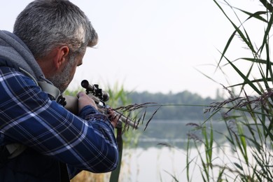 Man aiming with hunting rifle near lake outdoors. Space for text