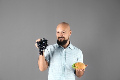 Overweight man with hamburger and grapes on gray background