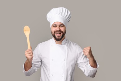 Photo of Happy young chef in uniform holding wooden spoon on grey background