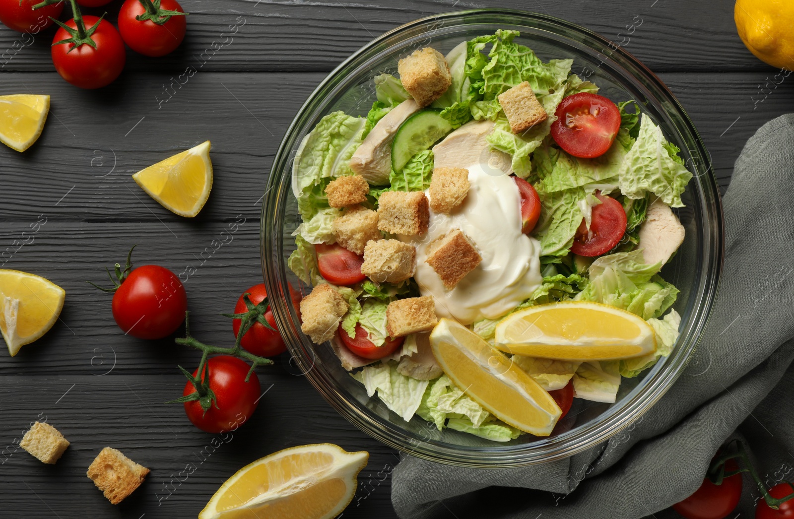 Photo of Bowl of delicious salad with Chinese cabbage, lemon, tomatoes and bread croutons on black wooden table, flat lay