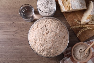 Fresh sourdough in bowl, bread, flour, water and spikes on wooden table, flat lay. Space for text