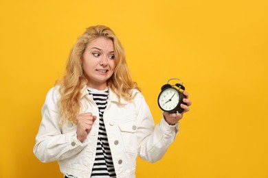 Emotional woman with alarm clock in turmoil over being late on yellow background