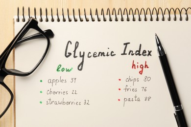 Photo of List with products of low and high glycemic index in notebook, glasses and pen on wooden table, top view