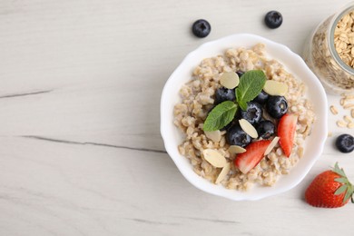 Photo of Tasty oatmeal with strawberries, blueberries and almond petals in bowl on white wooden table, flat lay. Space for text