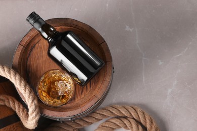 Photo of Whiskey with ice cubes in glass, bottle, wooden barrels and rope on grey marble table, top view. Space for text