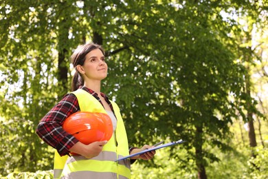 Forester with hard hat and clipboard examining plants in forest, space for text