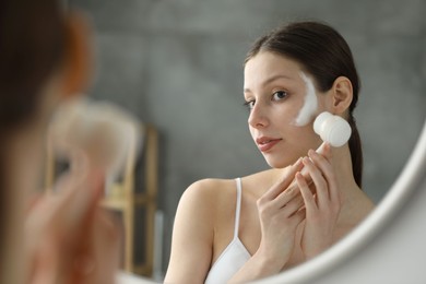 Photo of Young woman washing face with brush and cleansing foam near mirror in bathroom