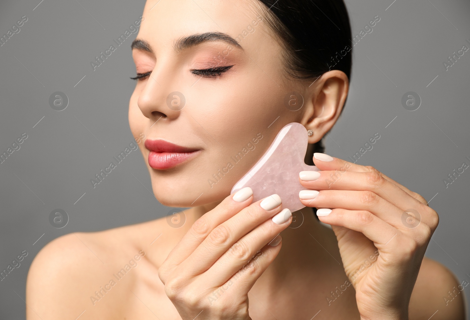 Photo of Beautiful young woman doing facial massage with gua sha tool on grey background, closeup