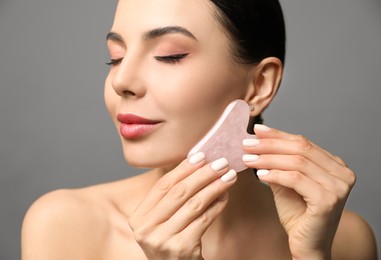 Beautiful young woman doing facial massage with gua sha tool on grey background, closeup