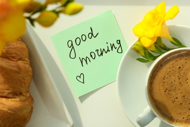 Photo of Cup of aromatic coffee with croissant, beautiful yellow freesia and Good Morning note on white table, flat lay