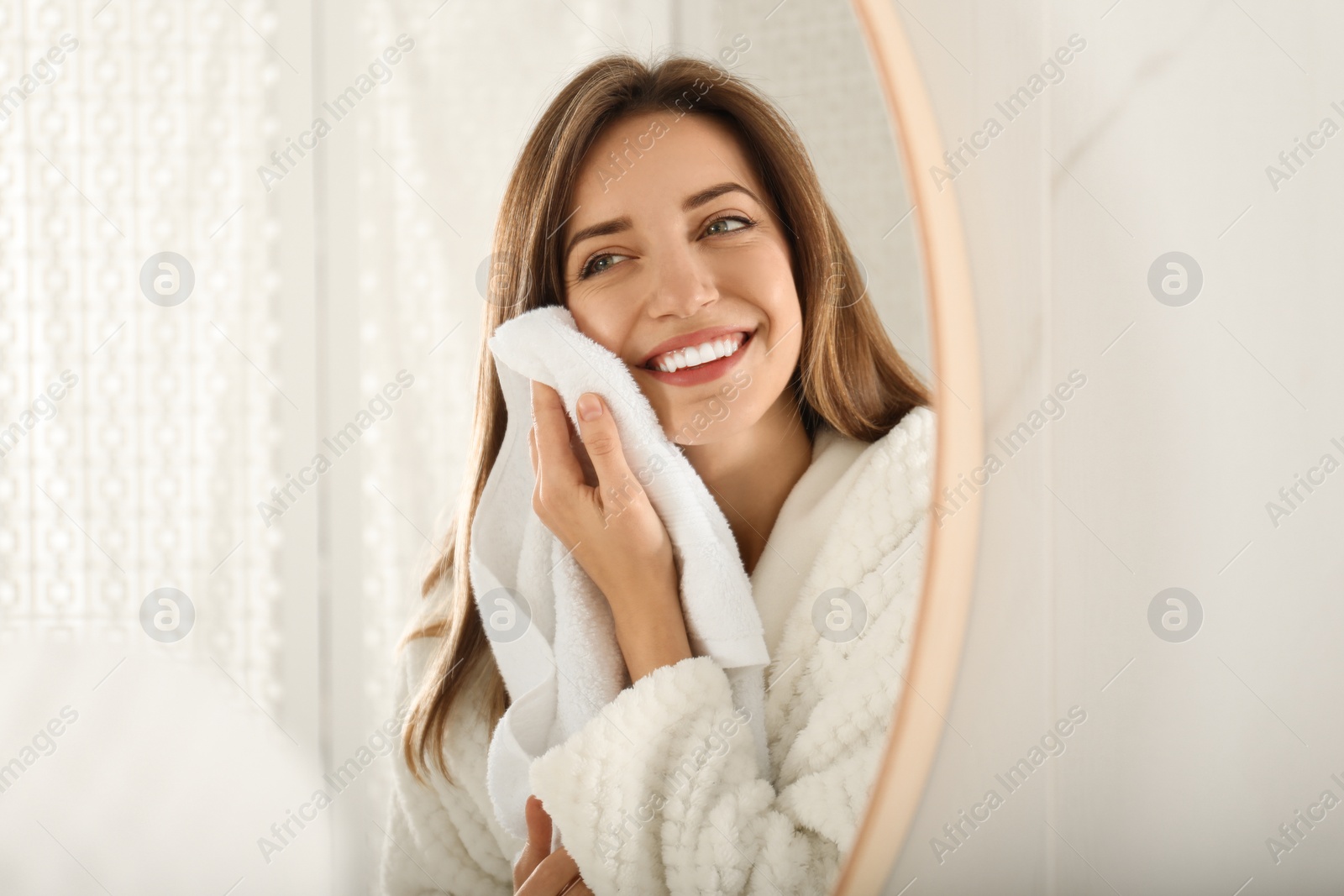 Photo of Young woman wiping face with towel near mirror in bathroom