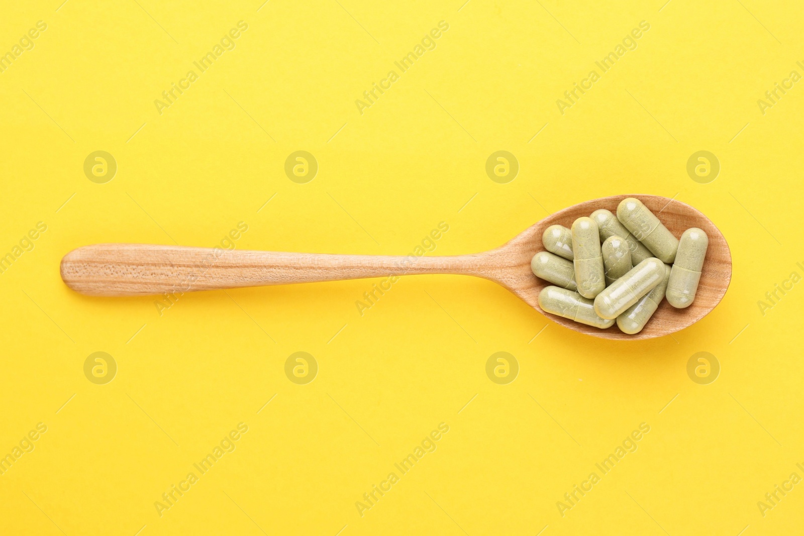 Photo of Vitamin pills in wooden spoon on yellow background, top view