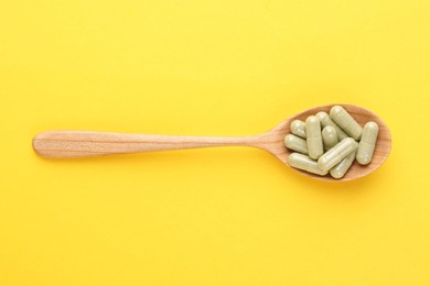 Vitamin pills in wooden spoon on yellow background, top view