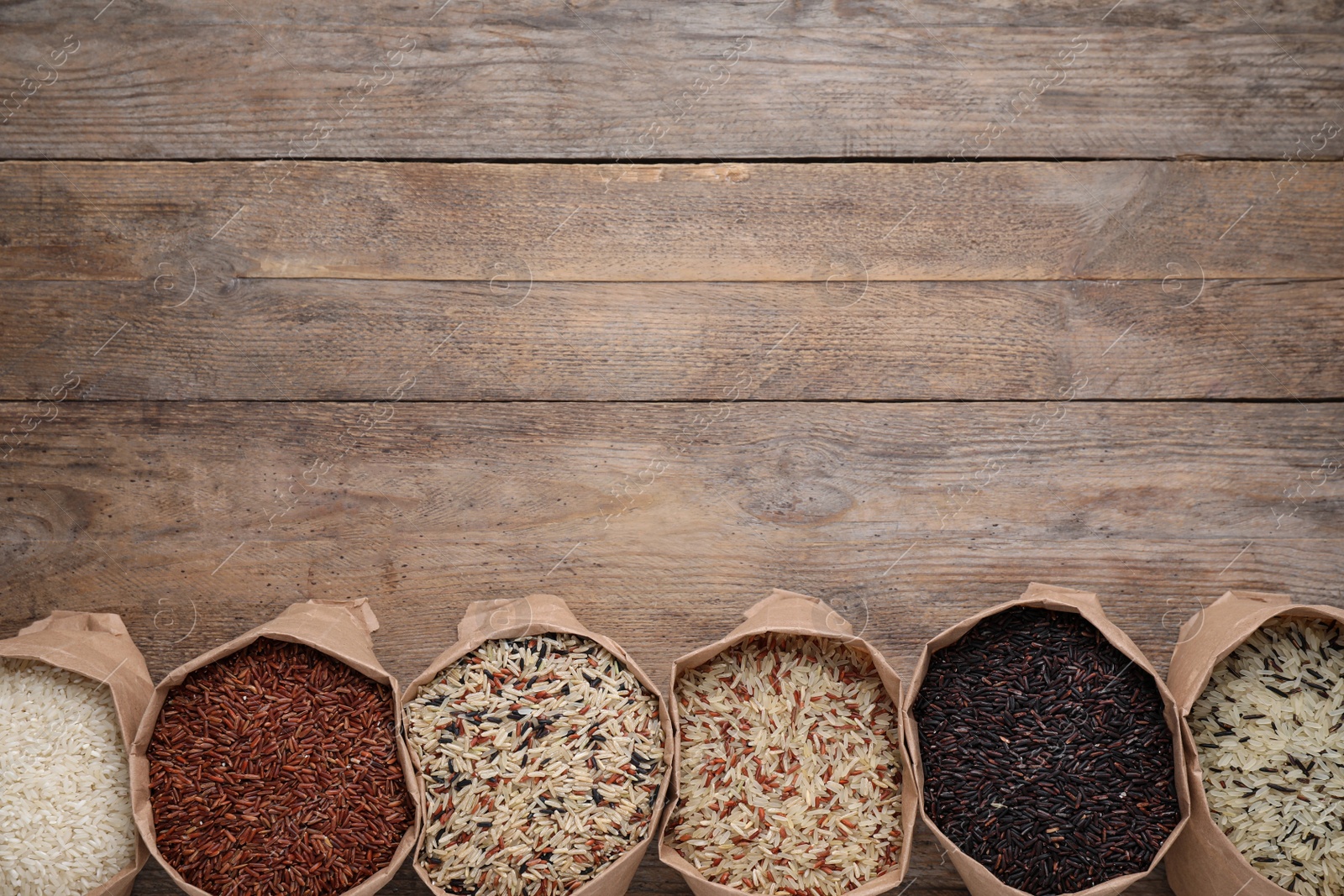 Photo of Different types of brown and polished rice in paper bags on wooden table, flat lay. Space for text