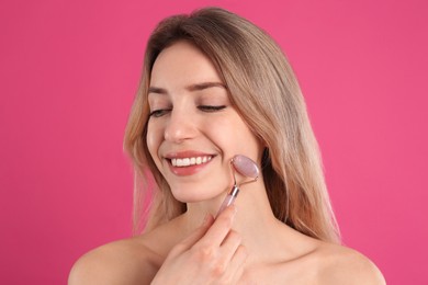 Young woman using natural rose quartz face roller on pink background