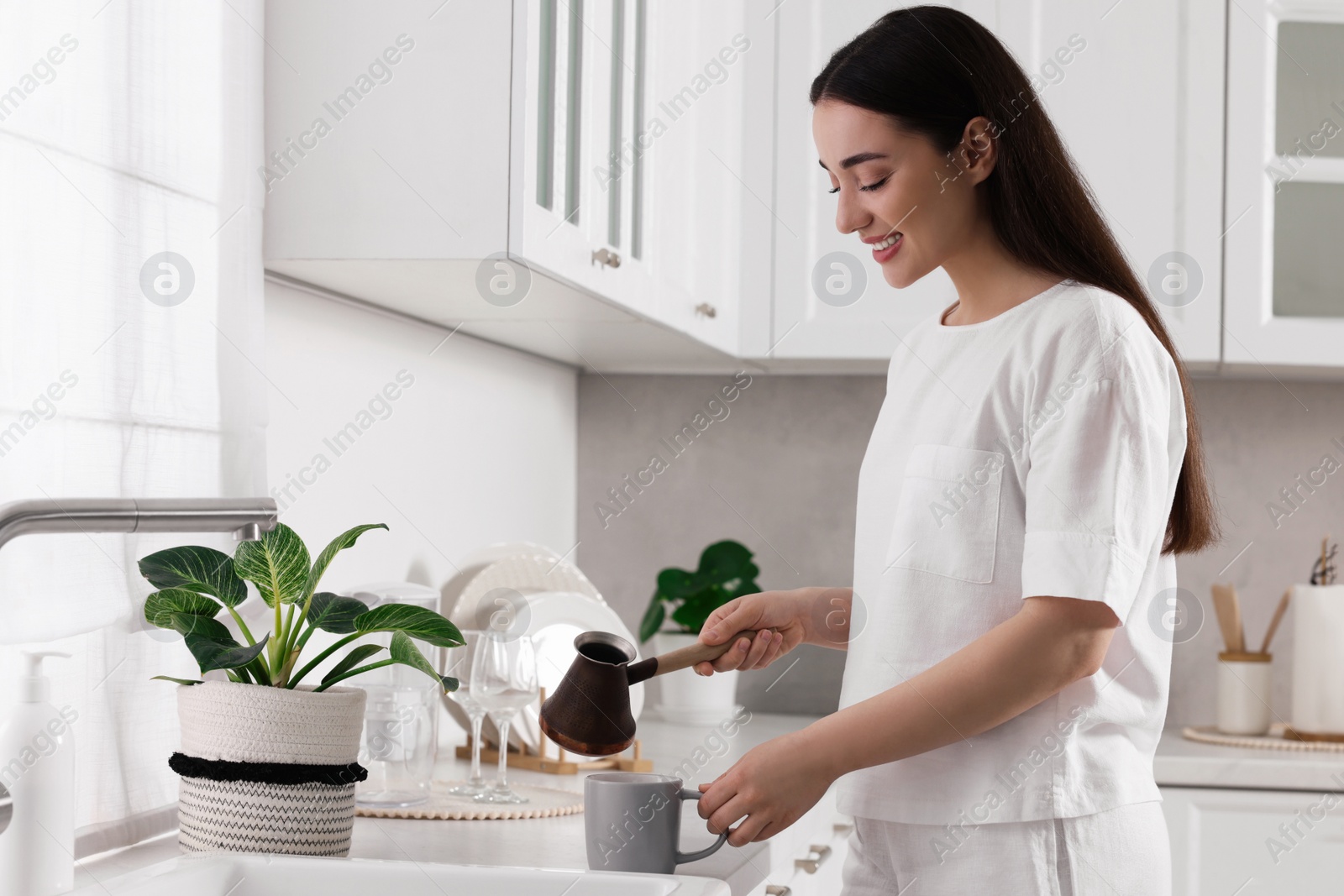 Photo of Beautiful woman pouring coffee from jezve into cup in kitchen. Lazy morning